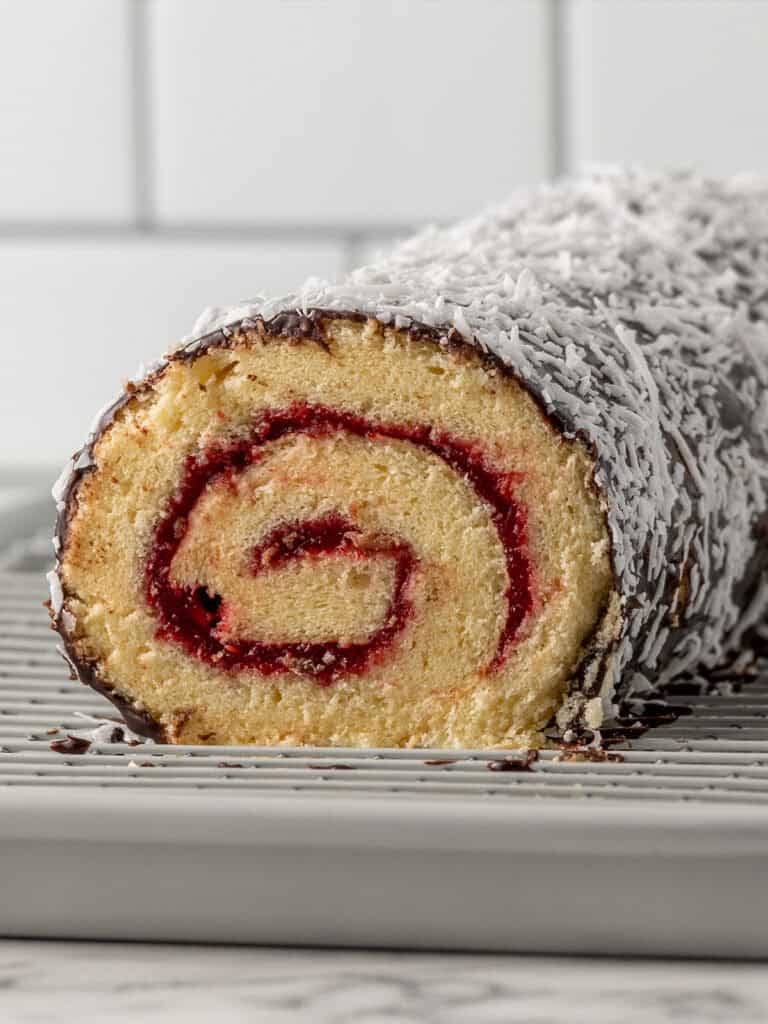 A close-up of a jam-filled Swiss roll cake covered in shredded coconut, displayed on a cooling rack with a white tiled background.