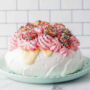 A pavlova topped with pink whipped cream and clusters of colorful sprinkles sits on a light green plate, against a white tiled background.