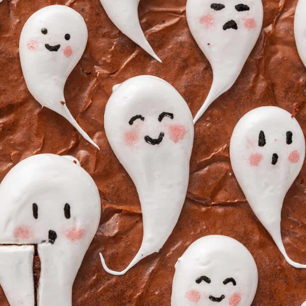 Several white meringue ghosts with hand-drawn faces and pink cheeks arranged on a chocolate brownie.