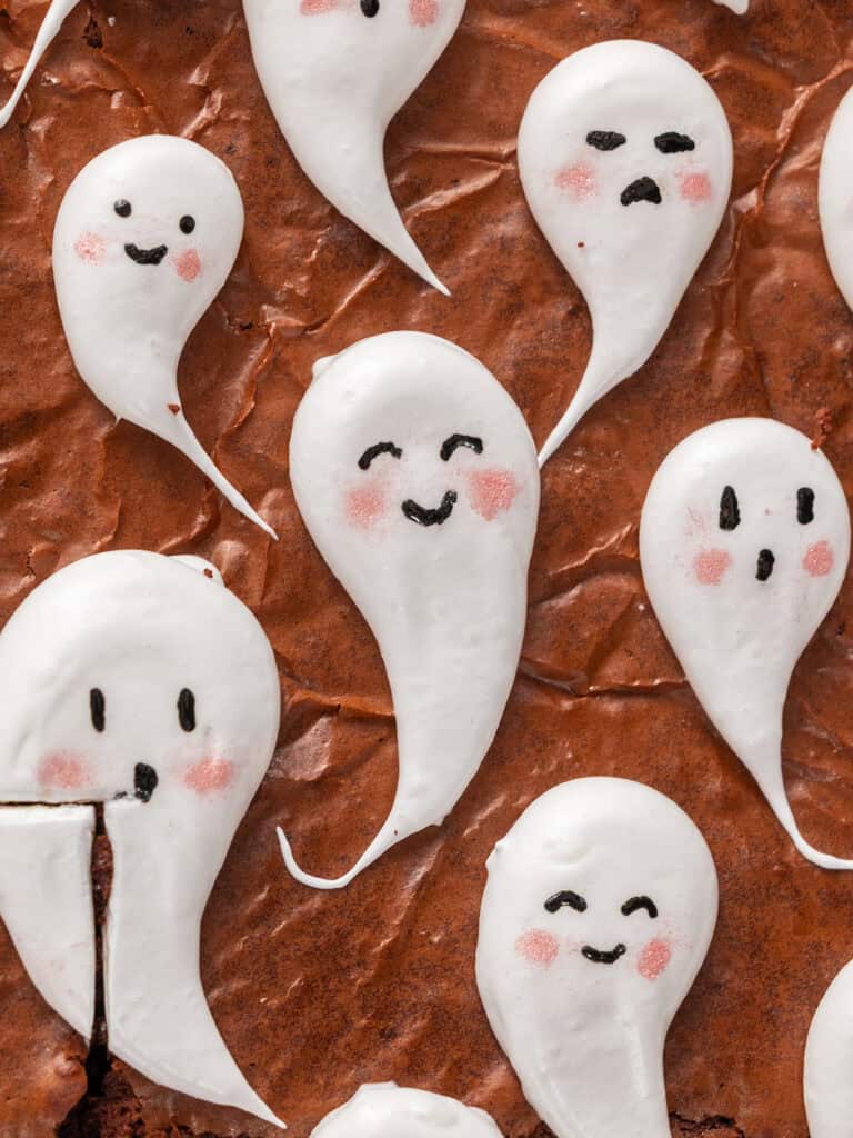 Brownie topped with several small white meringue ghosts, each decorated with hand-drawn faces and pink cheeks.