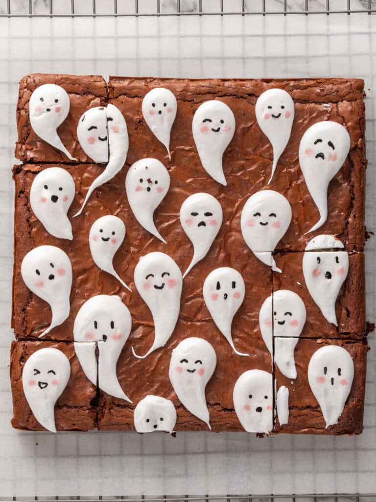 A tray of brownies cut into squares, each topped with a small, white meringue ghost with a hand-drawn face.