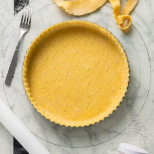 An unbaked pie crust in a round tart pan with fork marks, surrounded by a fork, rolling pin, dough scraps, and a sprinkle of flour on a marble surface.