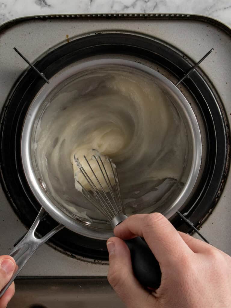 A person uses a whisk to stir a thick, flour and water mixture called tangzhong in a saucepan on a stovetop, likely making a roux or sauce.