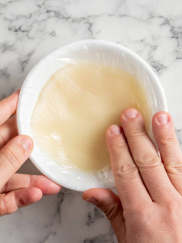 Two hands are covering a white bowl filled with tangzhong mixture using clear plastic wrap, set on a white marble surface.