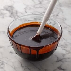 A glass bowl filled with glossy chocolate sauce, some dripping down the sides, sits on a marble surface. A white spatula rests in the bowl, coated with the sauce.