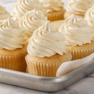 A metal tray filled with vanilla cupcakes topped with generous swirls of creamy white frosting, arranged neatly on a marble surface.