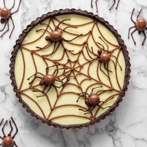 A tart with a chocolate crust and vanilla filling decorated with a chocolate spiderweb and candy spiders, surrounded by additional spider decorations on a marble surface.