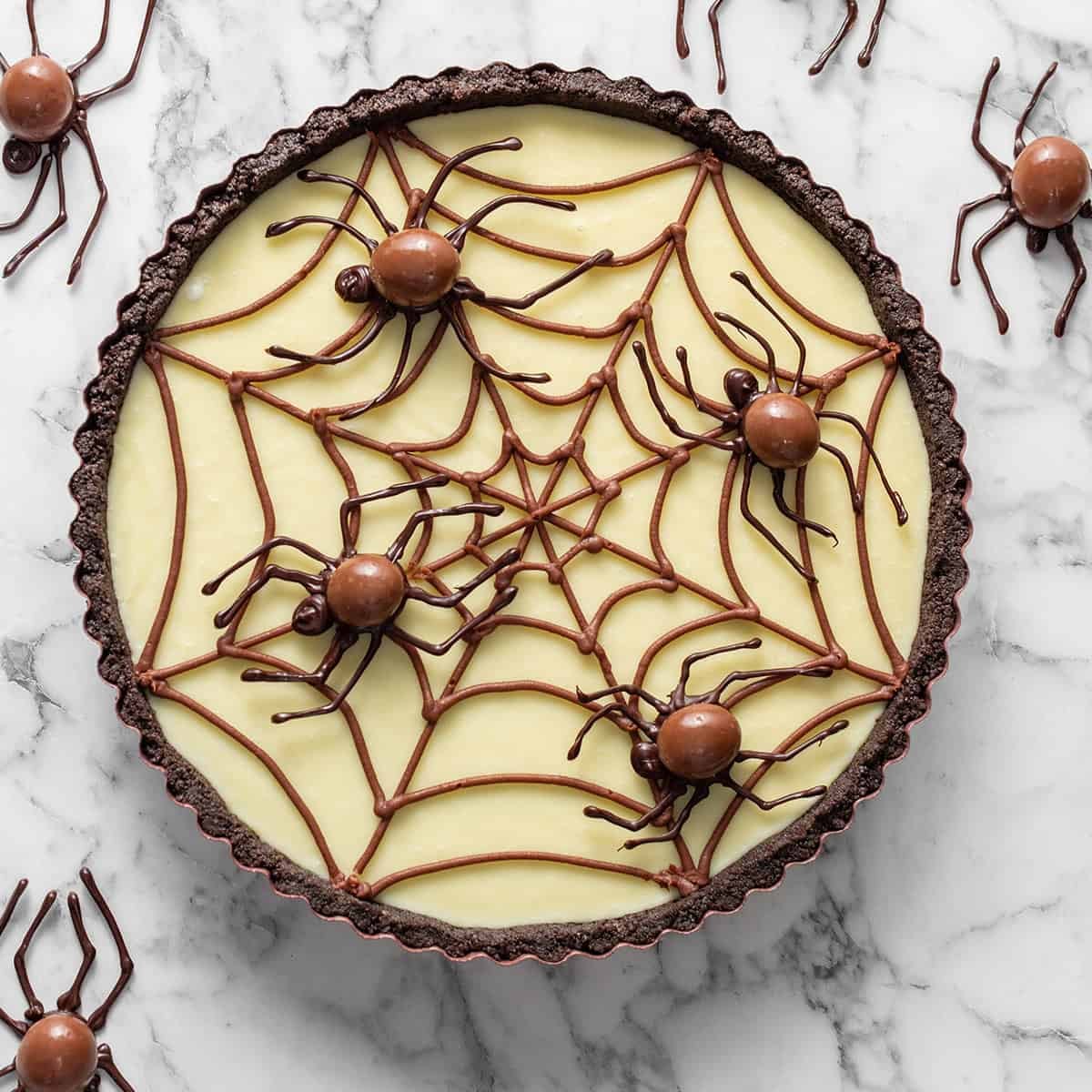A tart with a chocolate crust and vanilla filling decorated with a chocolate spiderweb and candy spiders, surrounded by additional spider decorations on a marble surface.
