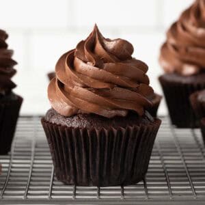 A chocolate cupcake with swirled chocolate frosting sits on a cooling rack, with more cupcakes in the background. The cupcake is in a dark brown paper liner.