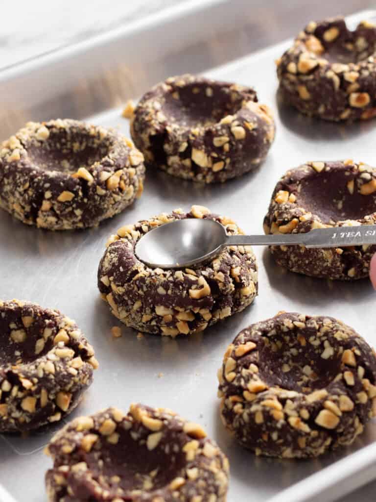 A metal teaspoon pressing an indentation into a chocolate cookie coated in crushed nuts on a baking sheet.