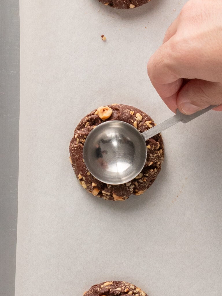 A hand uses a metal measuring spoon to press down on a chocolate hazelnut cookie on parchment paper.
