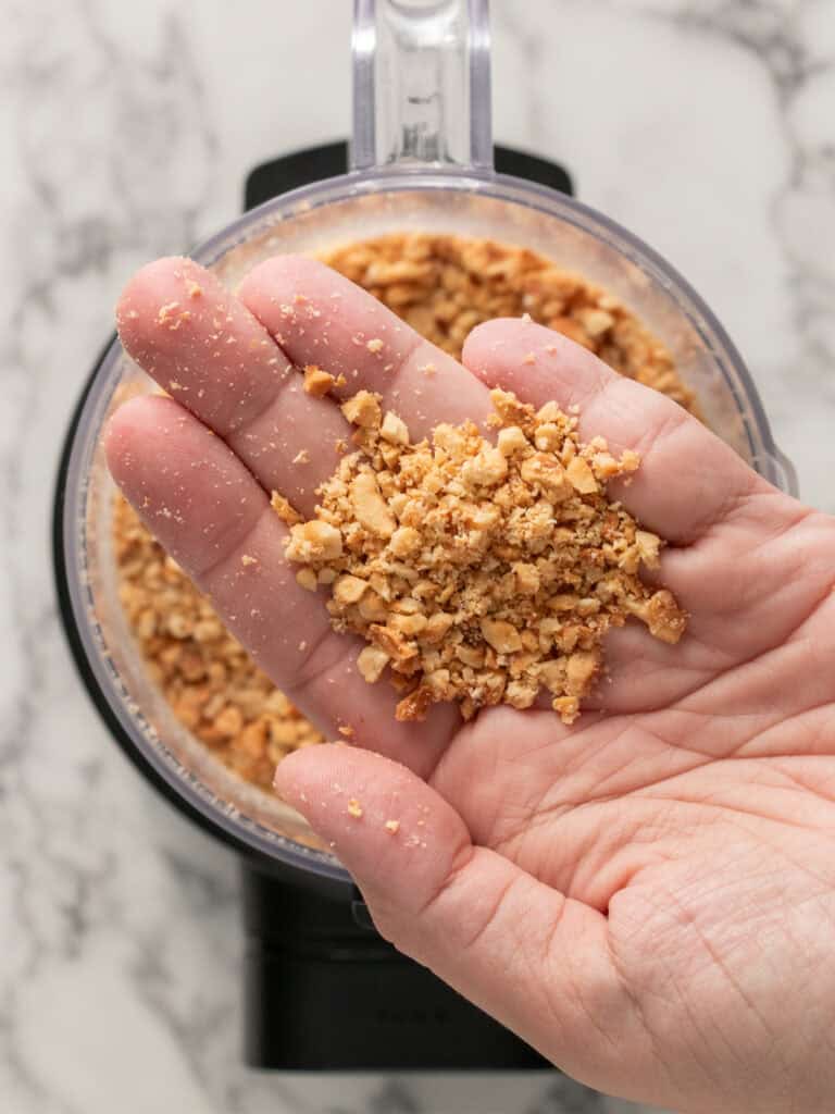 A hand holds a palmful of crushed nuts over an open food processor.