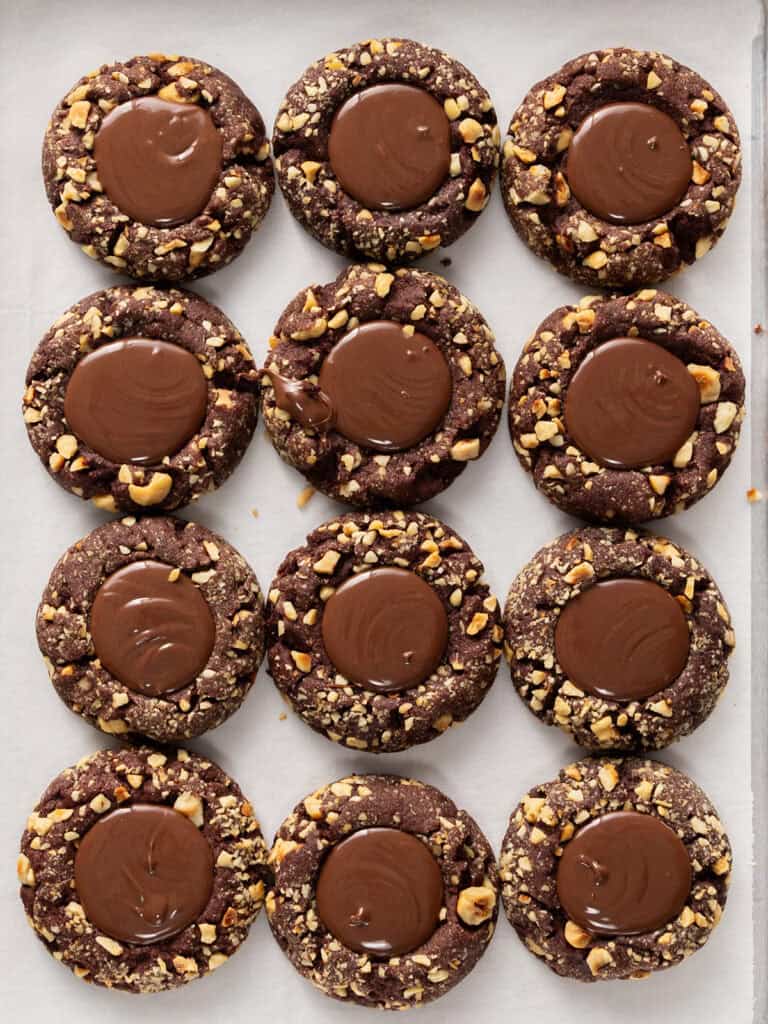 Twelve chocolate thumbprint cookies topped with chocolate ganache and chopped nuts, arranged on parchment paper.