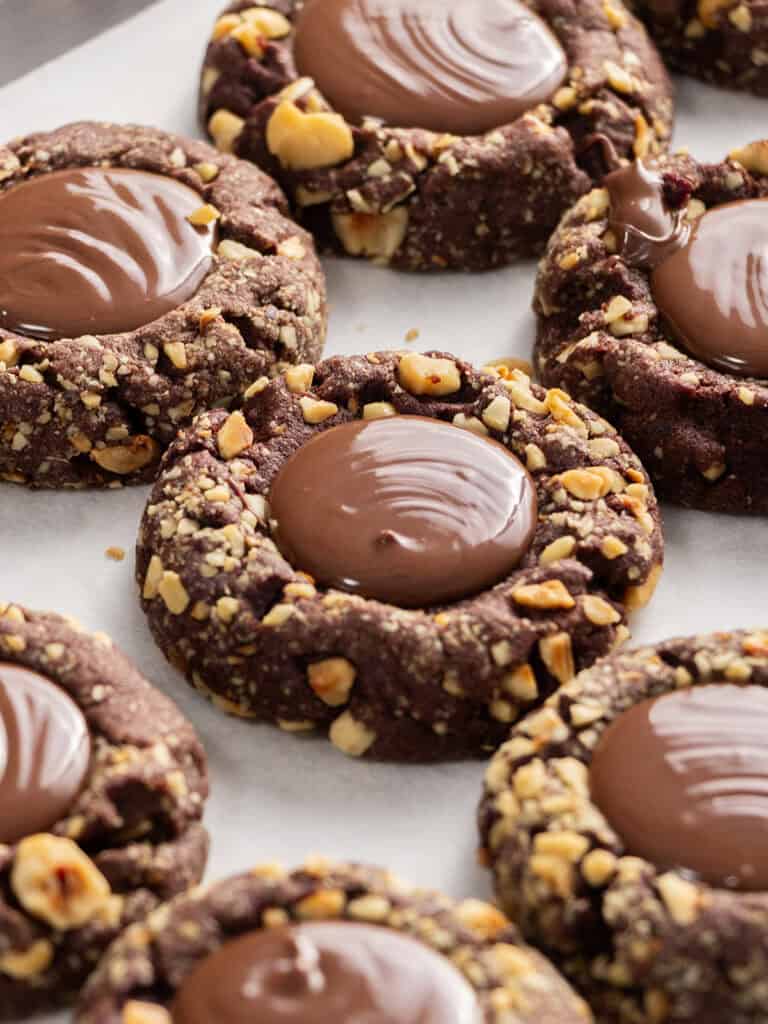 Chocolate thumbprint cookies topped with a dollop of chocolate ganache and rolled in chopped nuts, arranged on parchment paper.