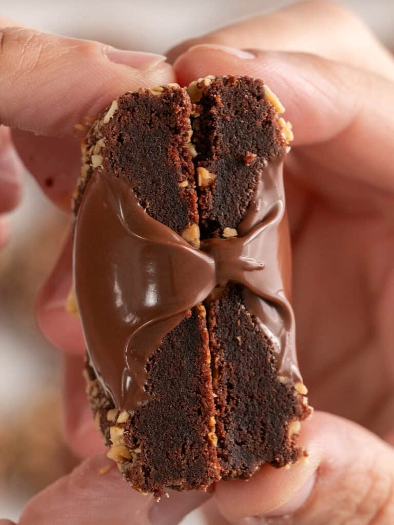 Two fingers hold a split chocolate brownie sandwich filled with melted chocolate and topped with nuts.