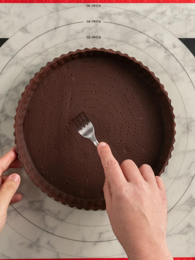 A hand uses a fork to prick holes in an unbaked chocolate tart crust set in a fluted pan, placed on a circular pastry mat with measurement guides.