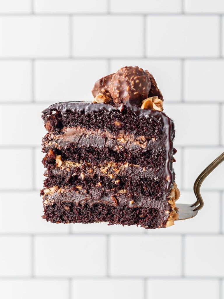 A single slice of chocolate hazelnut cake showing chocolate ganache filling and soft chocolate cake with chocolate candy on top on a spatula with a white background