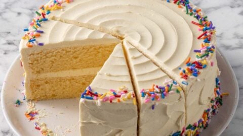 A round vanilla layer cake with white frosting and colorful sprinkles on the top edge and base. Several slices have been cut, revealing the soft yellow interior. The cake sits on a white plate on a marble surface.