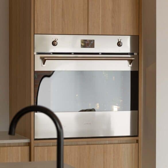 A square image side shot of an oven in an oven tower with soft glossy lighting and a black tap in the way set in a kitchen nook