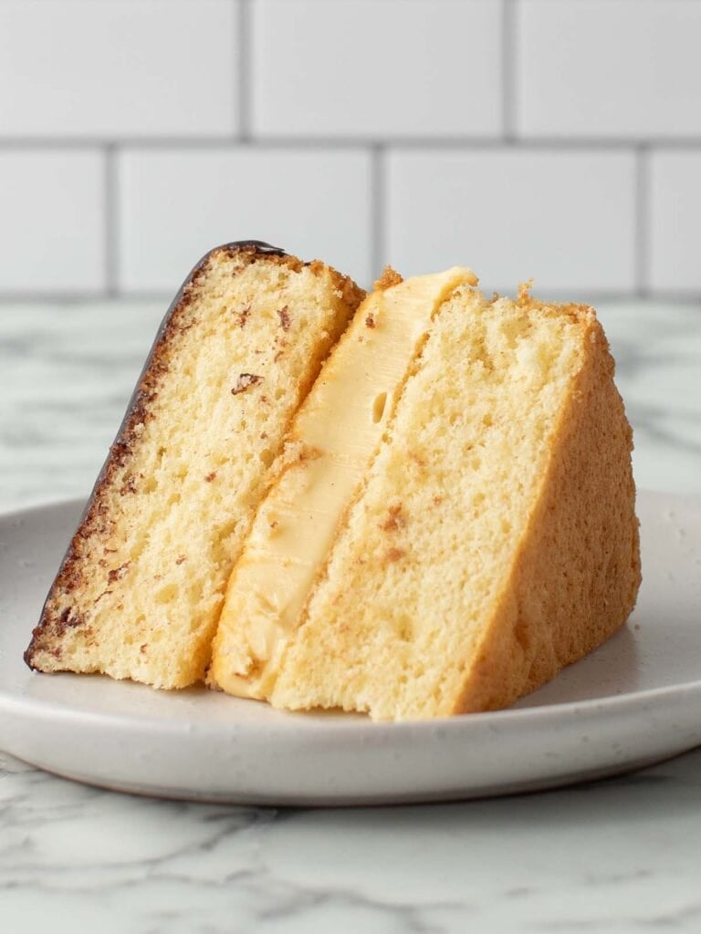 A single slice of vanilla sponge with custard in the middle and chocolate sauce on top sitting on its side on a plate