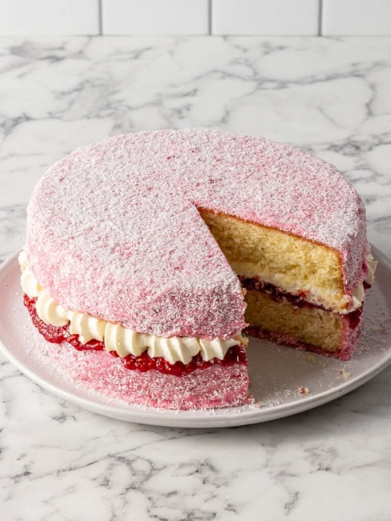 A pink covered sponge cake with jam and cream sitting a plate with a slice taken out