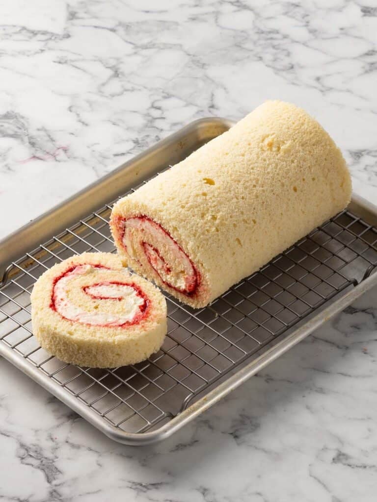 A spiral log cake filled with chantilly cream and raspberry jam sitting on a cooling rack with a slice sitting to one side