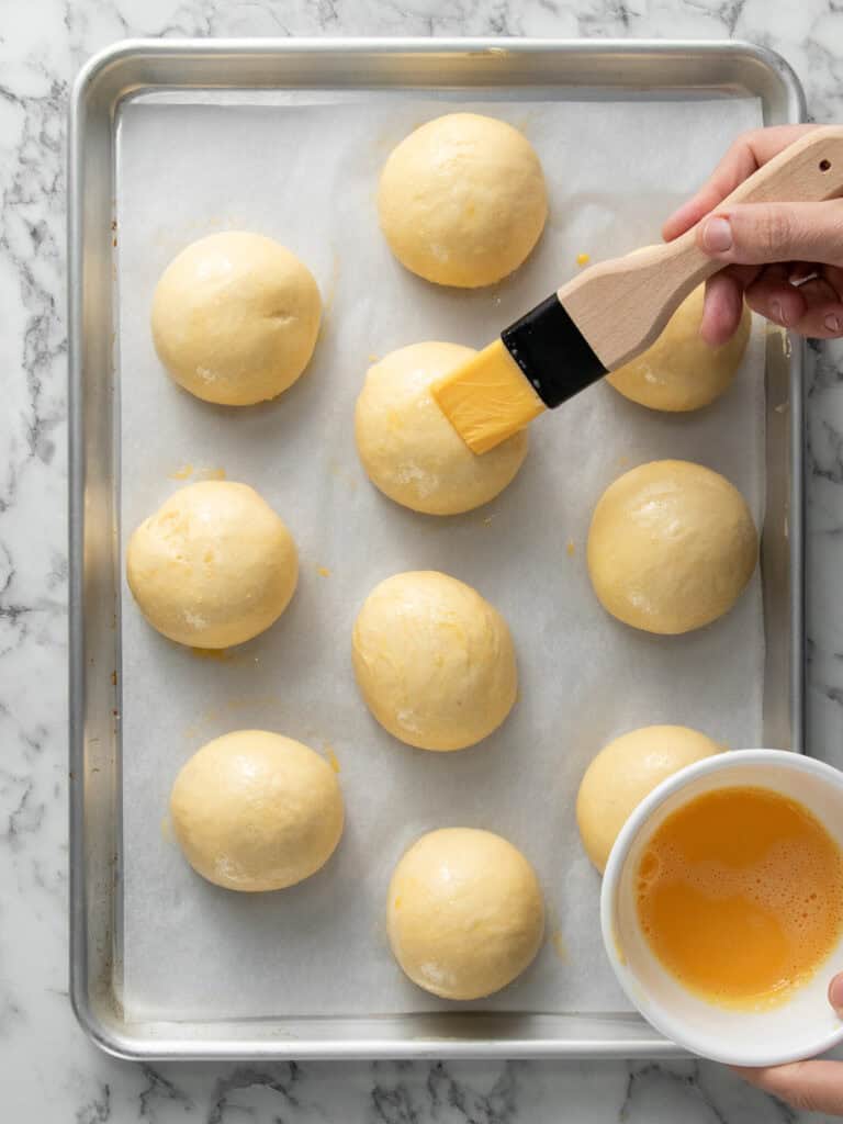 Brushing the risen buns with egg wash before baking