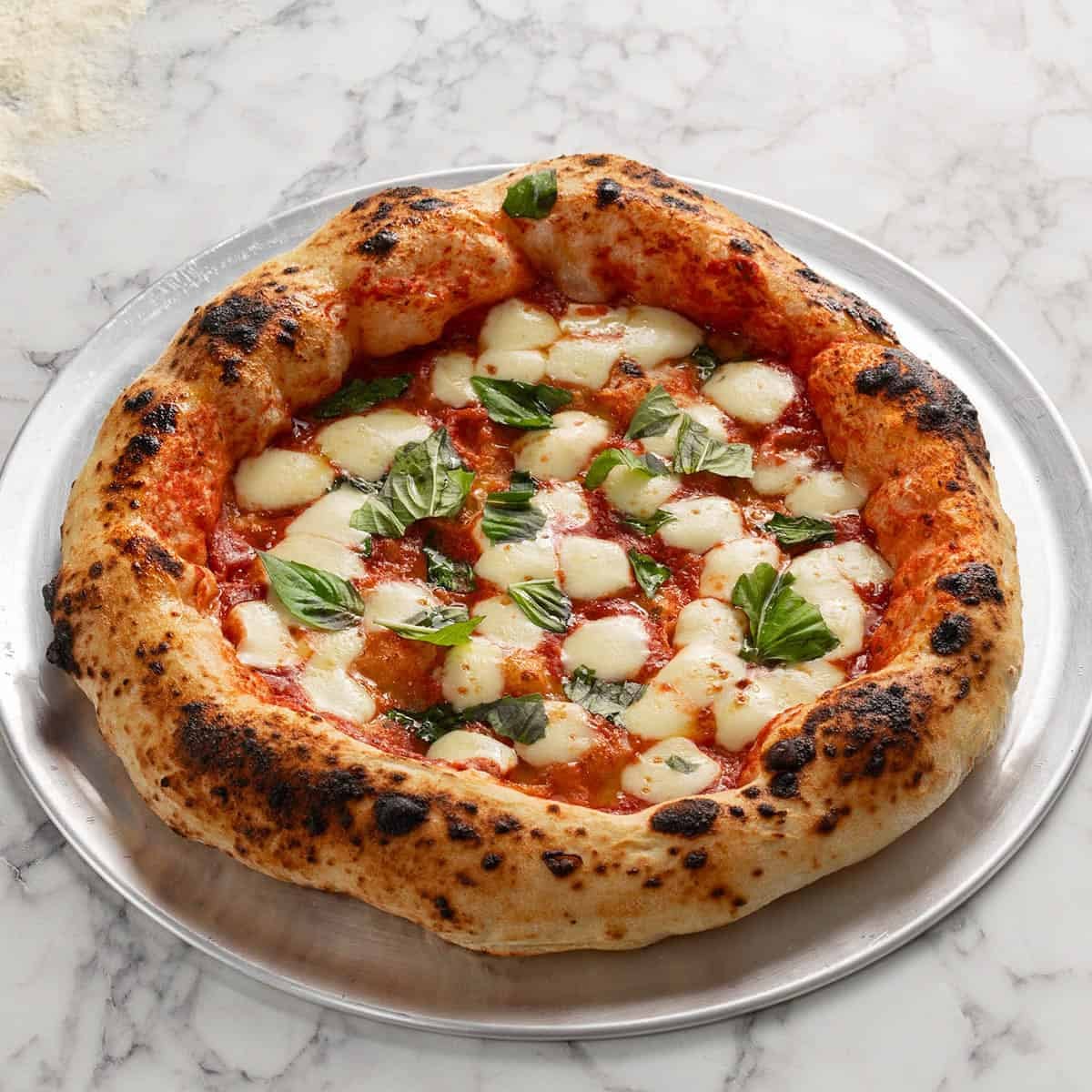 A Neapolitan-style pizza with bubbly, charred crust, melted mozzarella cheese, tomato sauce, and fresh basil leaves, served on a round metal tray atop a marble surface.