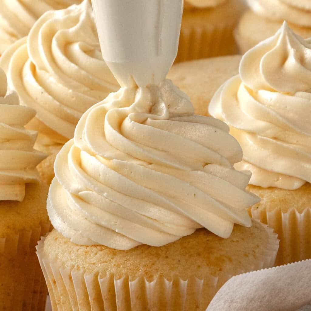 A close-up of vanilla cupcakes being frosted with a piping bag, swirling creamy white frosting on top of the cupcakes.