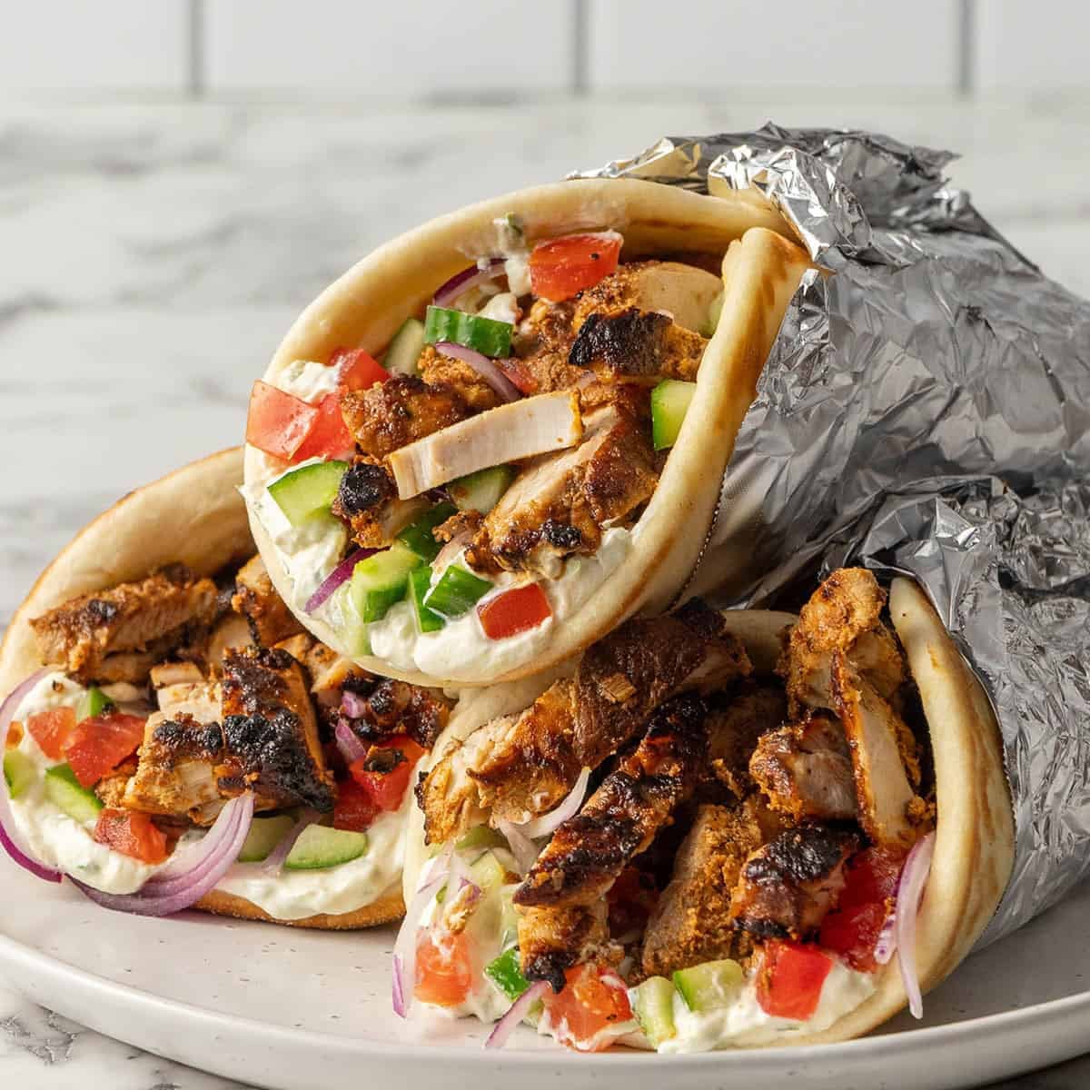 Three grilled chicken gyros wrapped in pita bread with tzatziki, diced tomatoes, cucumbers, and red onions, partially wrapped in foil, are stacked on a plate against a marble background.