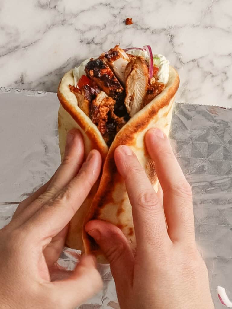 Two hands are holding a pita wrap filled with grilled chicken, sliced red onions, and sauce, over a piece of foil on a marble surface.