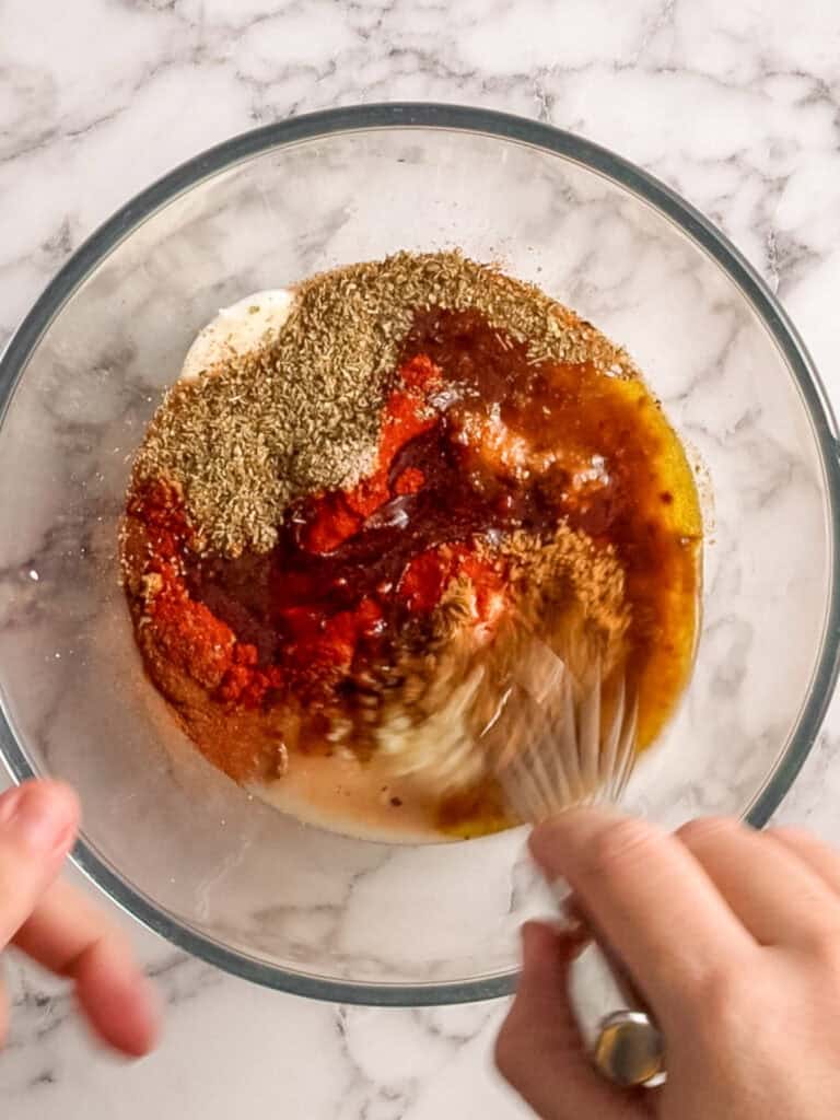 A hand uses a whisk to mix various spices, oil, and creamy ingredients in a glass bowl on a marble countertop.
