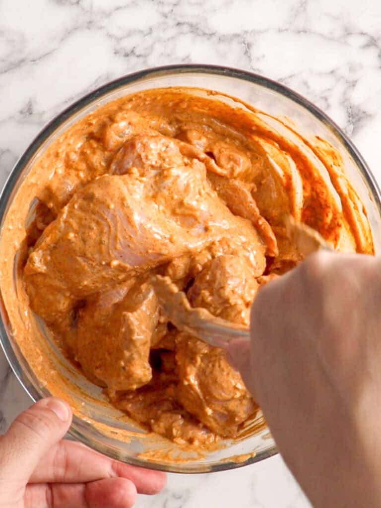 A hand is mixing raw chicken pieces with a thick, orange-brown marinade in a glass bowl on a marble countertop.