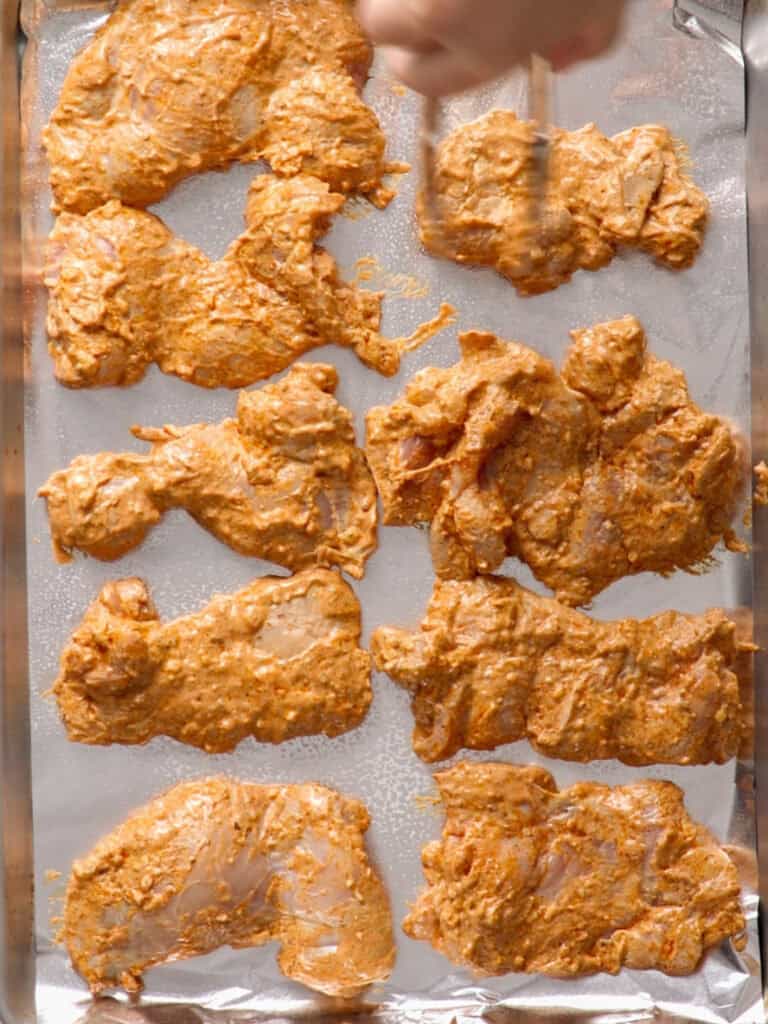 A hand arranges pieces of marinated raw chicken coated in a reddish-orange spice mixture on a foil-lined baking sheet.
