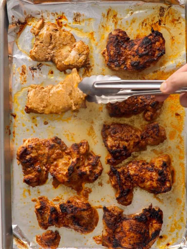 A hand uses metal tongs to place a piece of marinated, cooked chicken onto a foil-lined baking sheet with several other pieces of browned, roasted chicken.