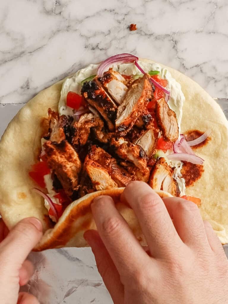Two hands folding a flatbread wrap filled with grilled chicken, sliced red onions, diced tomatoes, and a creamy white sauce on a marble surface.