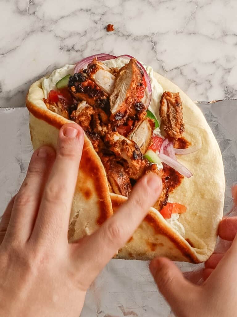 A person folds a pita bread filled with grilled meat, sliced red onions, tomatoes, and lettuce on a marble surface.