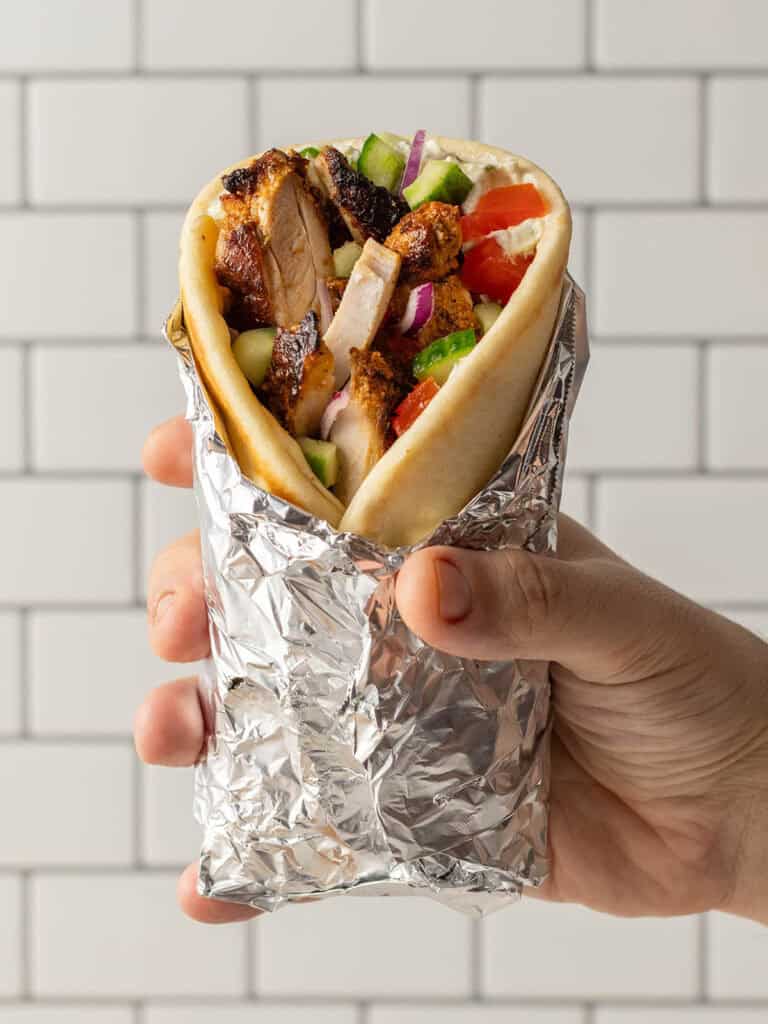 A hand holding a wrapped pita sandwich filled with grilled chicken, diced tomatoes, cucumbers, red onions, and sauce, against a white tiled background.