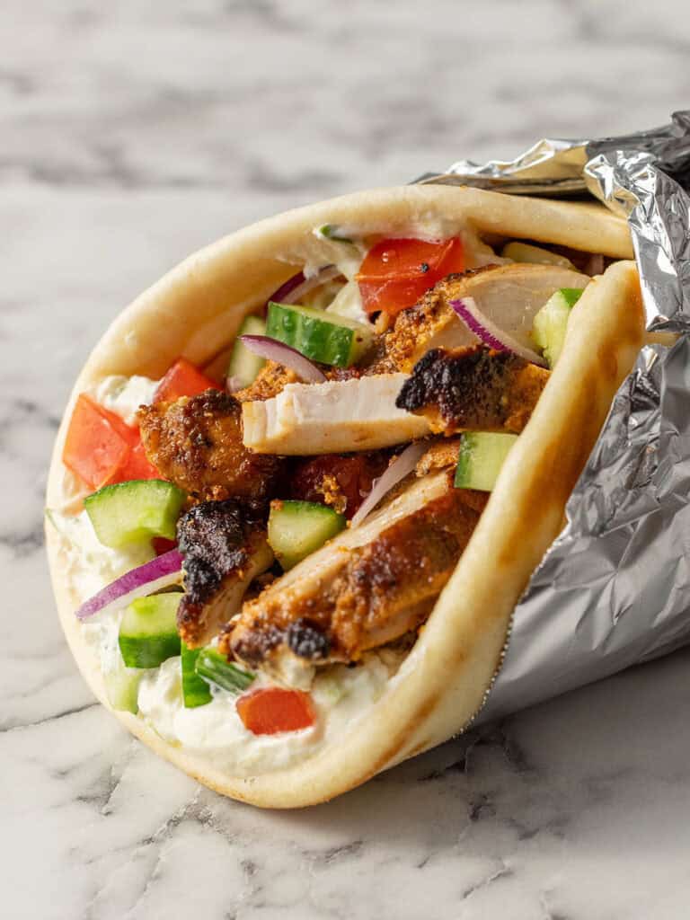 A close-up of a chicken gyro wrapped in foil, filled with grilled chicken, cucumber, tomato, red onion, and tzatziki sauce, on a marble surface.