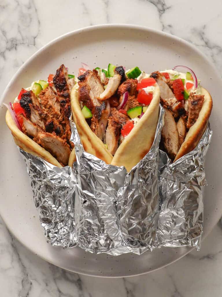 Three gyros wrapped in foil, filled with grilled chicken, chopped tomatoes, cucumbers, onions, and sauce, served on pita bread, displayed on a white marble surface.