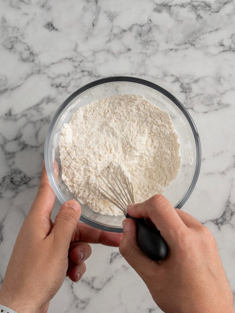A person holding a glass bowl of flour and mixing it with a metal whisk on a white marble countertop.