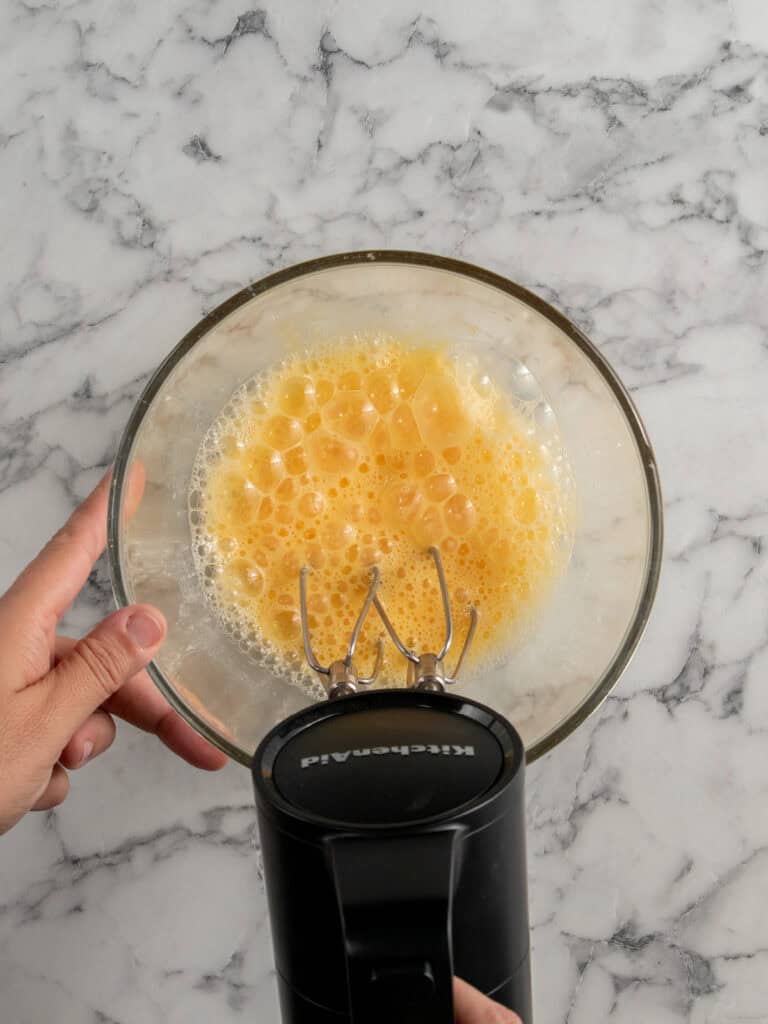 A hand holds a glass bowl filled with beaten eggs, while using a black KitchenAid hand mixer on a white marble countertop.