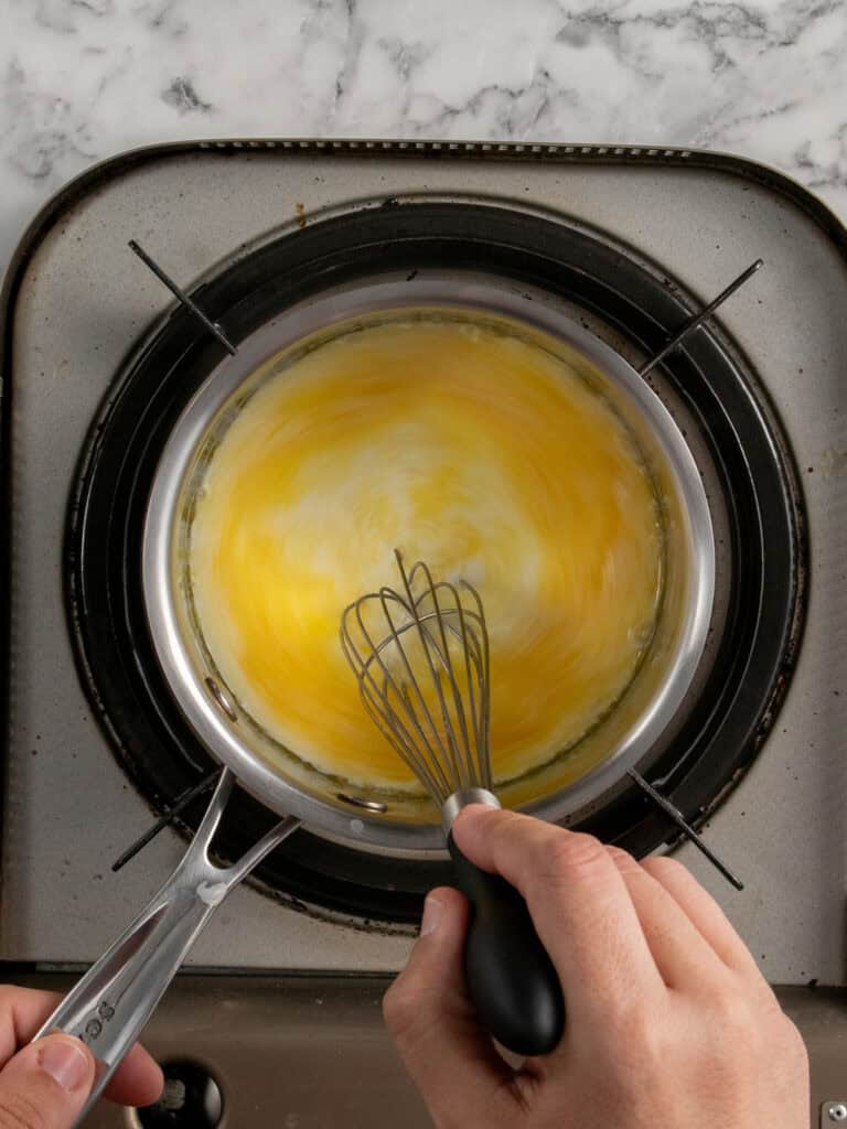 A person whisking eggs in a metal pan on a stovetop, creating a swirling yellow mixture. The stovetop has a marble countertop in the background.