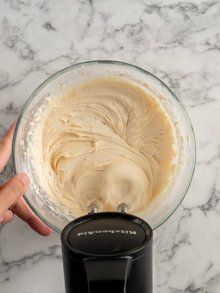 A hand holds a glass bowl of creamy, light-colored batter being mixed with a black electric hand mixer on a marble countertop.