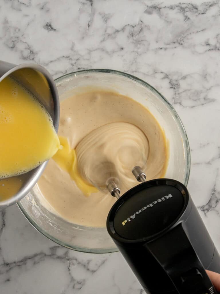 A hand mixer blends batter in a glass bowl while a yellow liquid is poured in from a metal saucepan, all set on a white marble countertop.