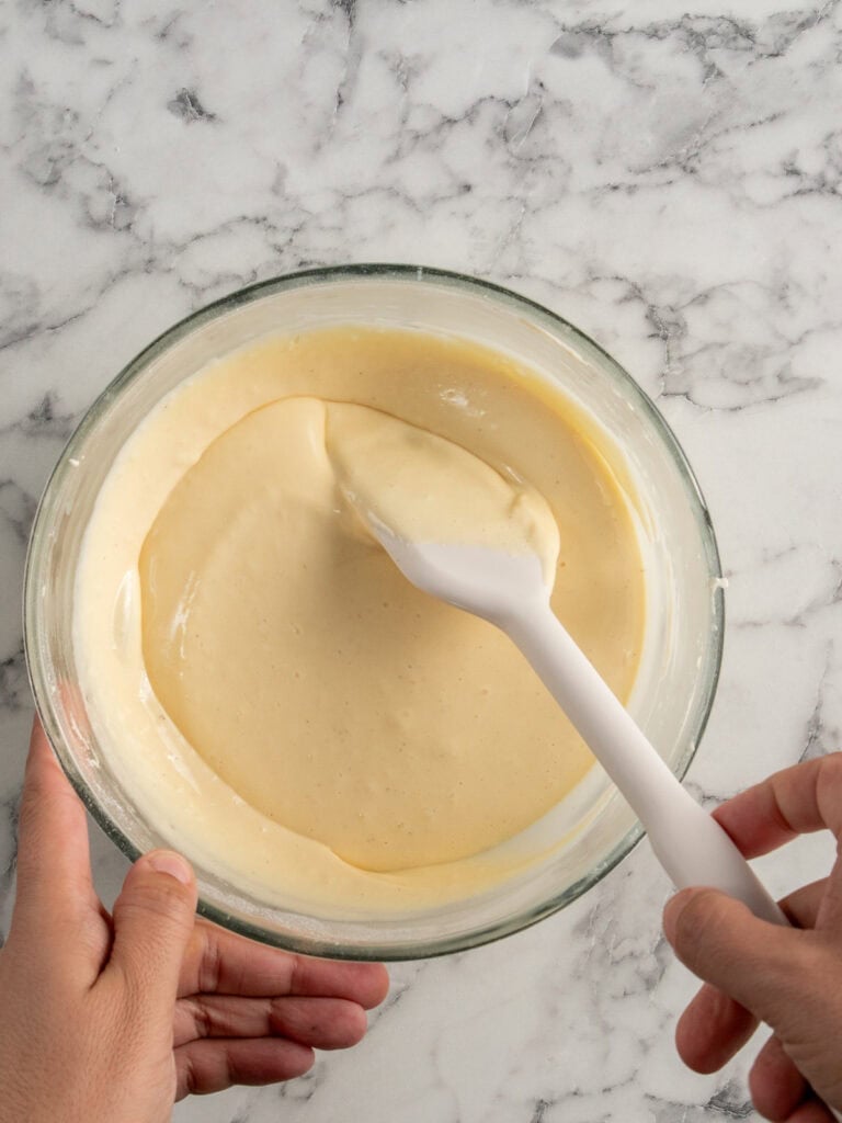 A person holds a glass bowl of smooth, pale batter on a marble surface, stirring it with a white spatula.