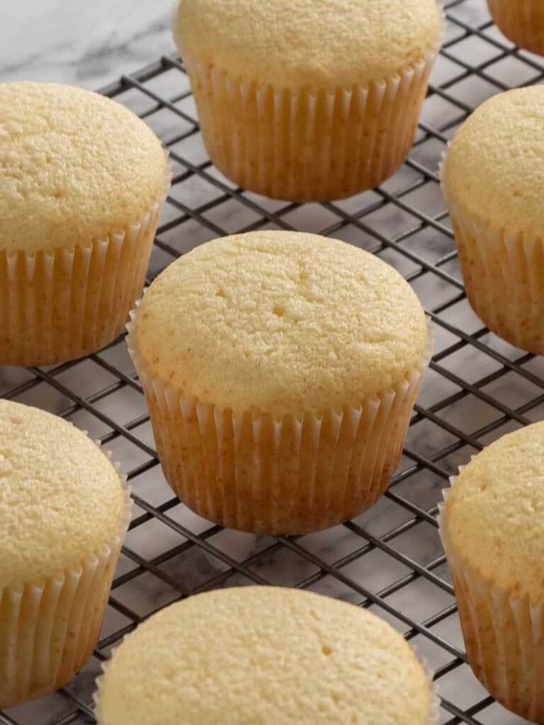 Freshly baked plain cupcakes in white paper liners are cooling on a metal wire rack. The cupcakes are golden and evenly spaced on the rack, which sits on a light-colored marble surface.
