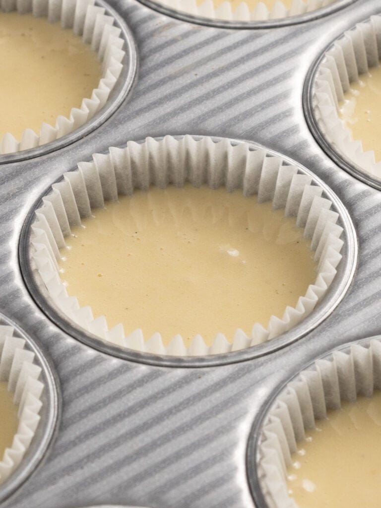 Close-up of a muffin tin lined with white paper cupcake liners, filled with smooth, pale yellow batter, ready to be baked.