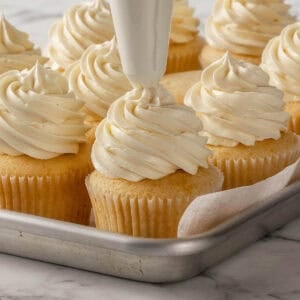 A piping bag adds swirls of creamy frosting onto a vanilla cupcake, surrounded by several similarly decorated cupcakes on a metal baking tray.