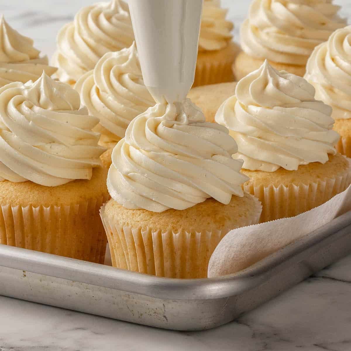 A piping bag adds swirls of creamy frosting onto a vanilla cupcake, surrounded by several similarly decorated cupcakes on a metal baking tray.